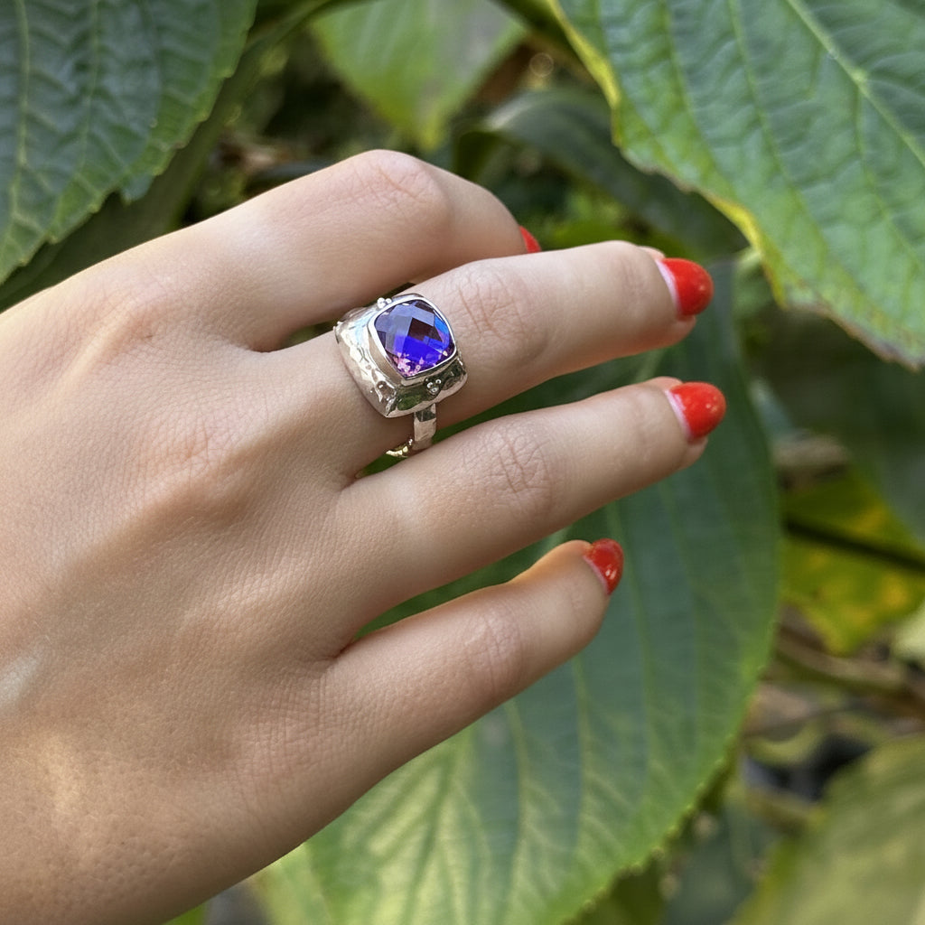 A hand with red nail polish wearing a silver ring with a square-cut deep blue gemstone, set against a background of green leaves.