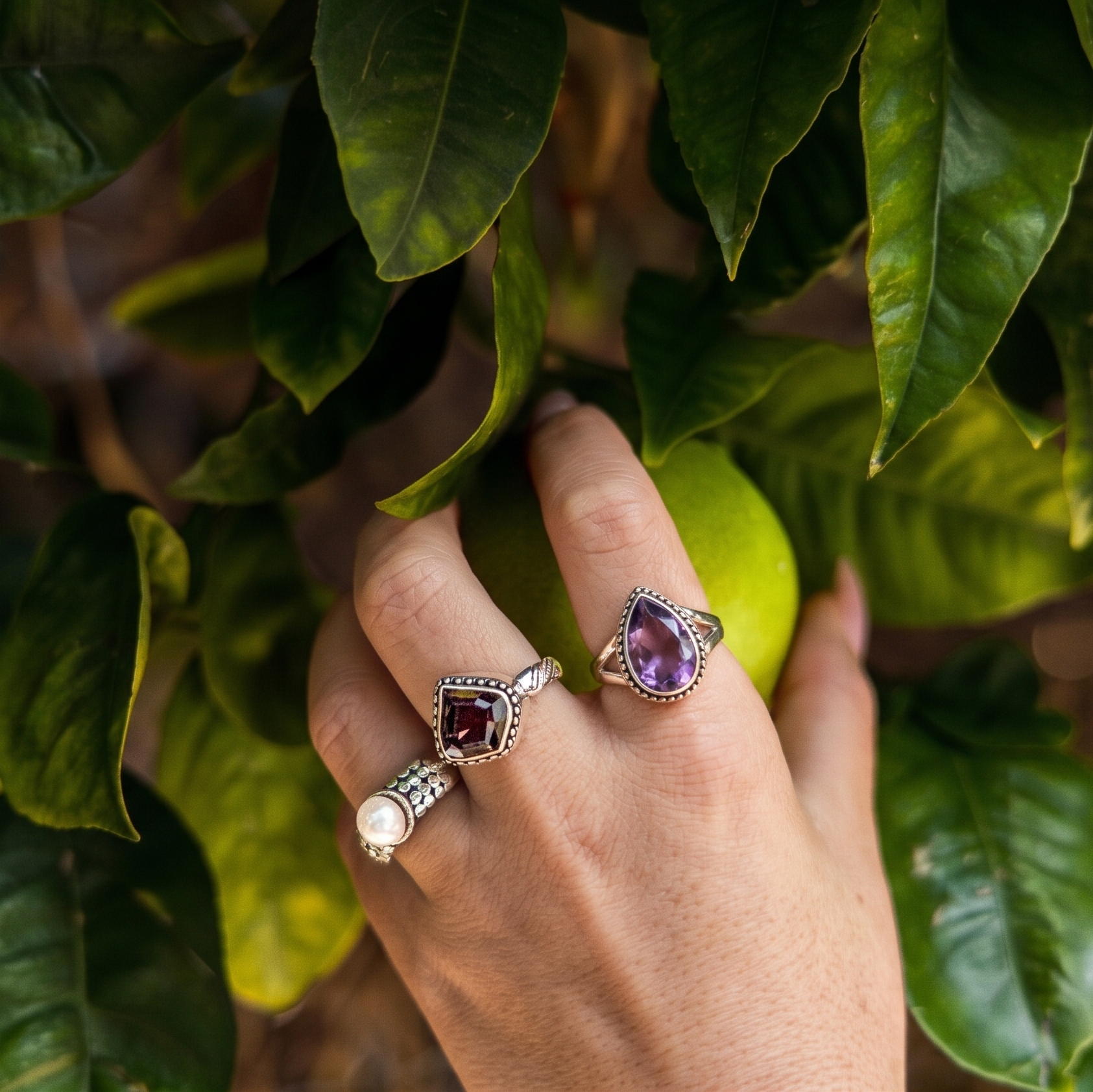 Hand wearing three rings with gemstones against a leafy background