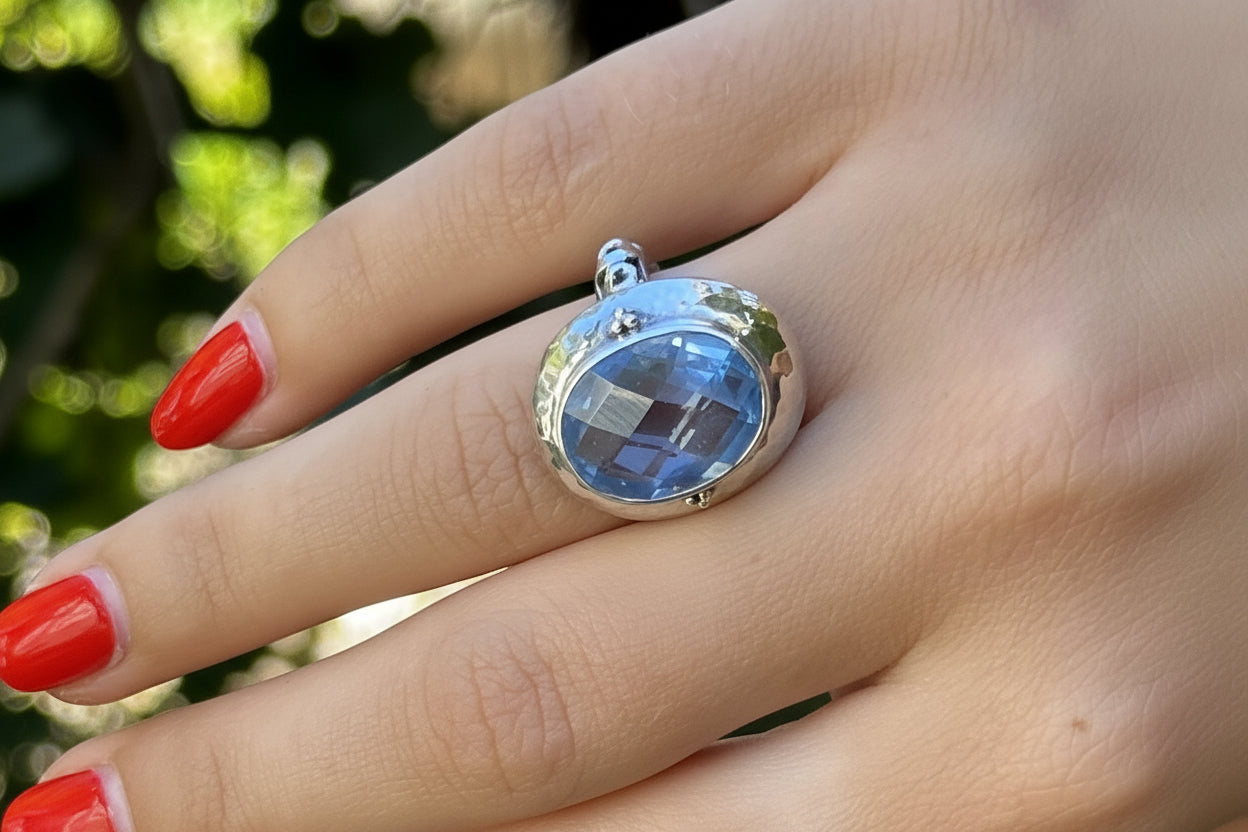 A close-up of a hand with red nail polish wearing a silver ring featuring a faceted oval blue gemstone, set against a blurred green outdoor background.