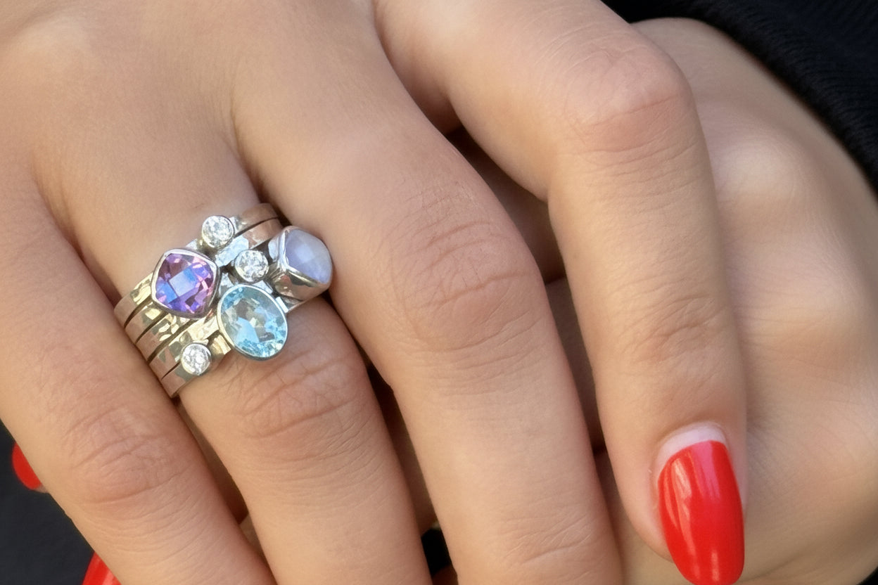 A close-up of a hand wearing a cluster ring with multiple gemstones, including a prominent light blue marquise-cut stone, a purple heart-shaped stone, a pale lavender oval stone, and small clear accent stones on a silver-toned band.